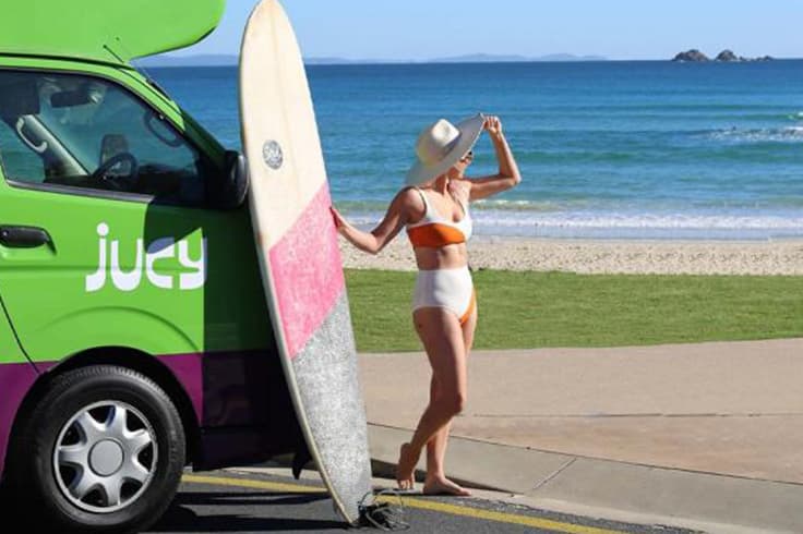 road trip itinerary brisbane to byron bay 1 Girl at the beach with a surfboard and a JUCY campervan