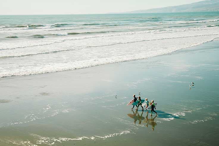 Take A Kiwi Road Trip This November Surfers walking on beach down to water